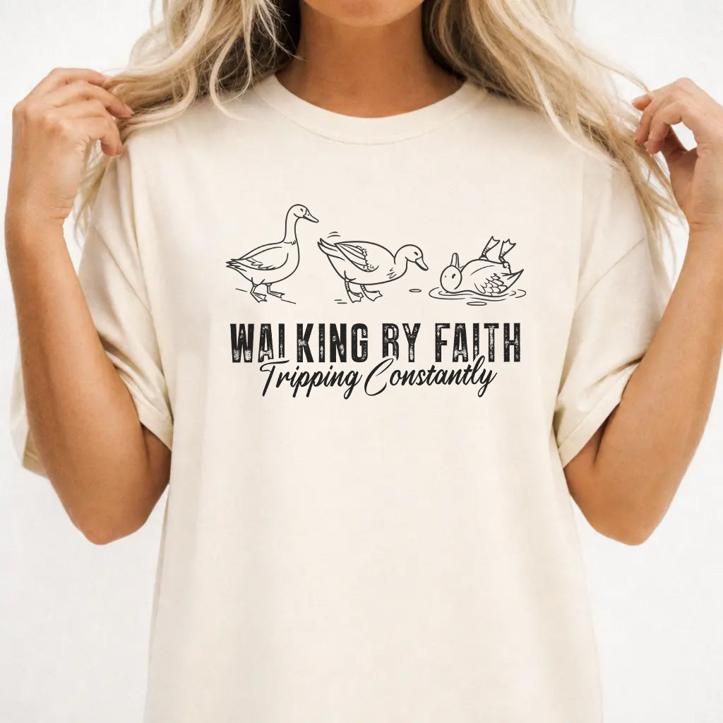 Woman wearing an ivory shirt with three duck illustrations and the phrase Walking by Faith Tripping Constantly on the front