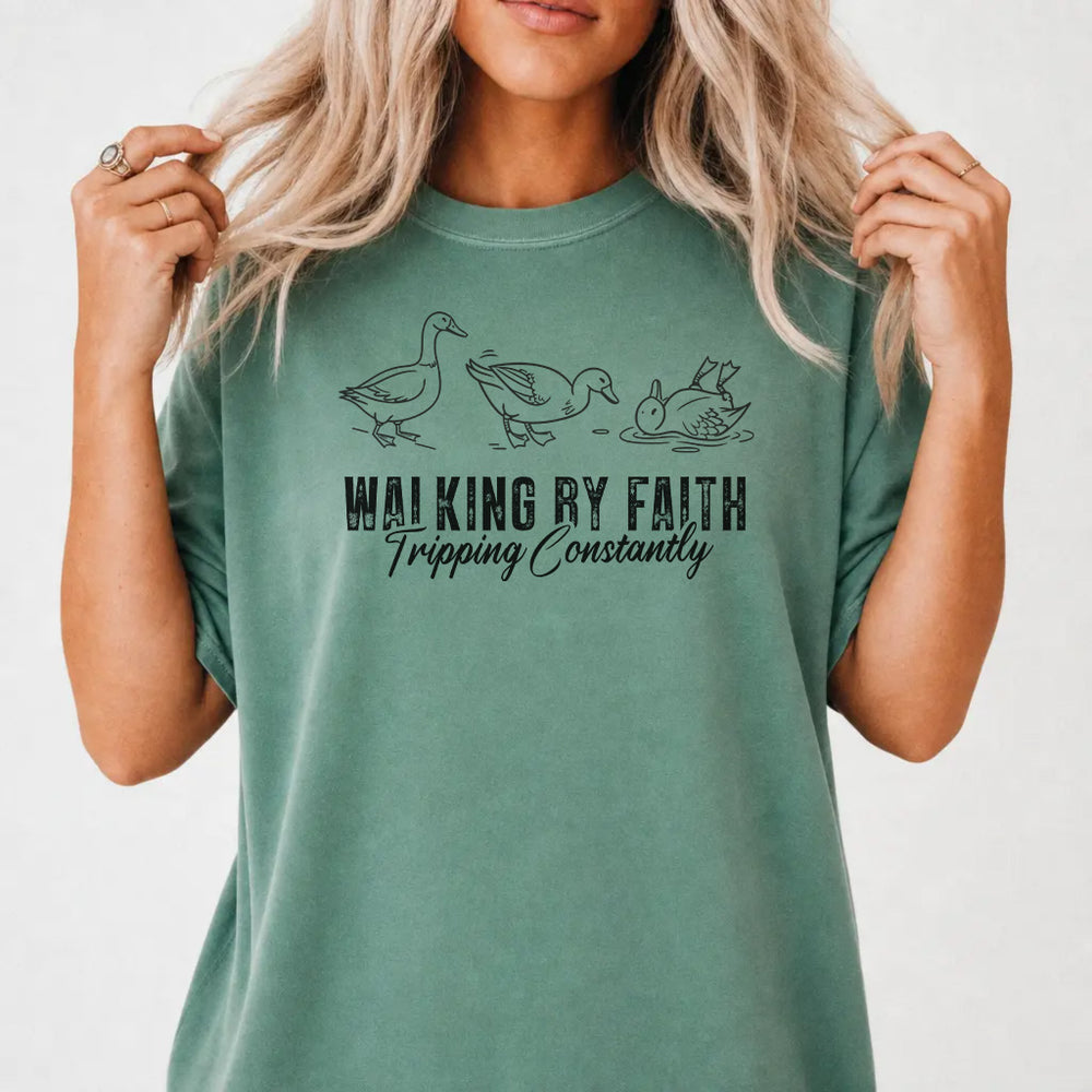 Woman wearing a light green shirt with three duck illustrations and the phrase Walking by Faith Tripping Constantly on the front