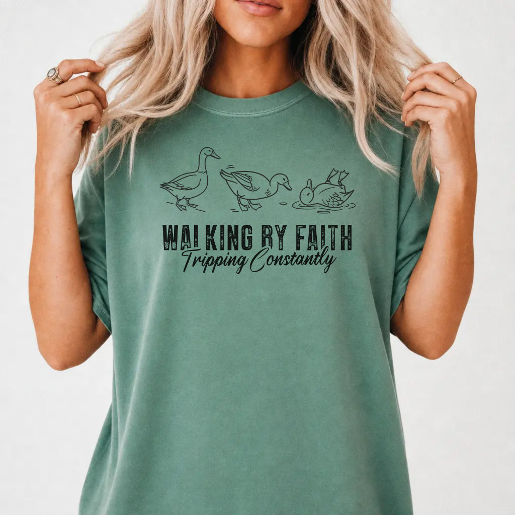 Woman wearing a light green shirt with three duck illustrations and the phrase Walking by Faith Tripping Constantly on the front