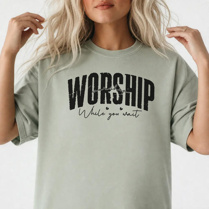 Woman wearing a bay shirt with bold distressed lettering and script text that reads Worship While You Wait on the front