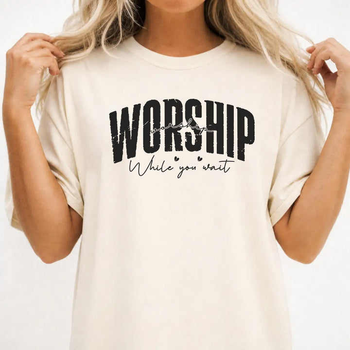Woman wearing an ivory shirt with bold distressed lettering and script text that reads Worship While You Wait on the front