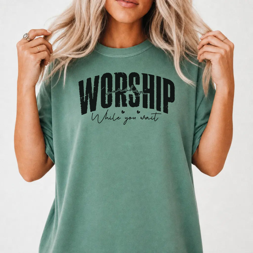 Woman wearing a light green shirt with bold distressed lettering and script text that reads Worship While You Wait on the front