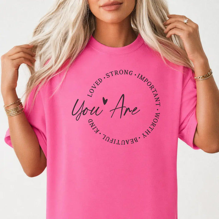 Woman wearing a neon pink shirt with a circle of affirmation words including loved strong worthy and beautiful around the script phrase You Are on the front