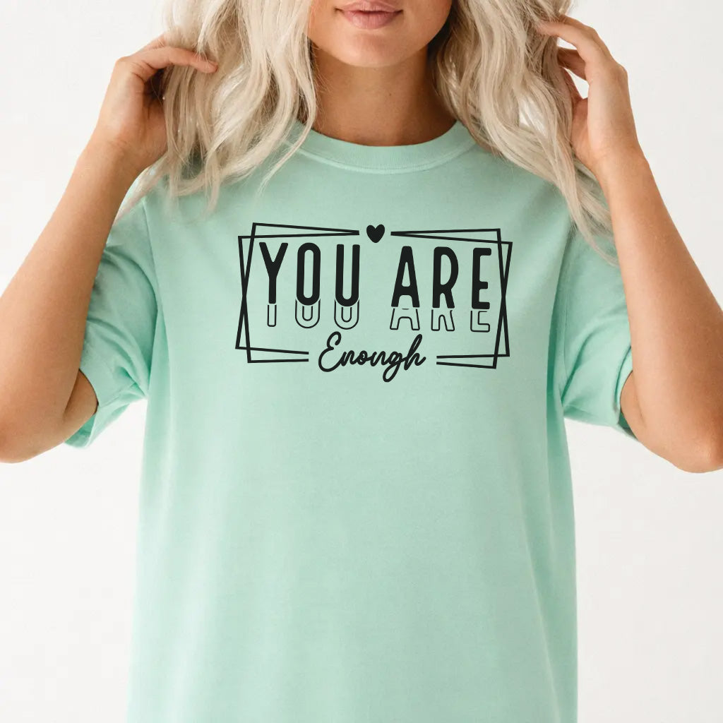 Woman wearing an island reef graphic tee with bold stacked lettering and a heart accent that reads You Are Enough on the front