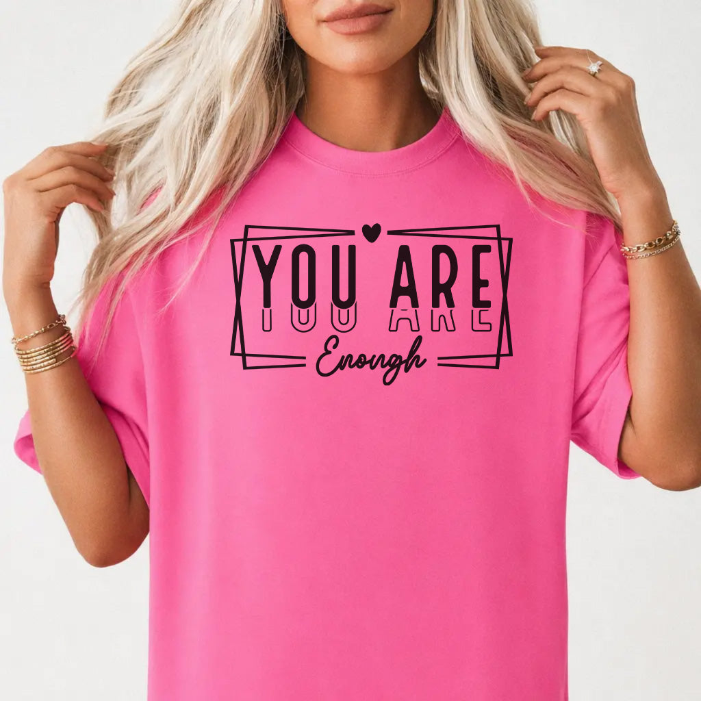 Woman wearing a neon pink graphic tee with bold stacked lettering and a heart accent that reads You Are Enough on the front