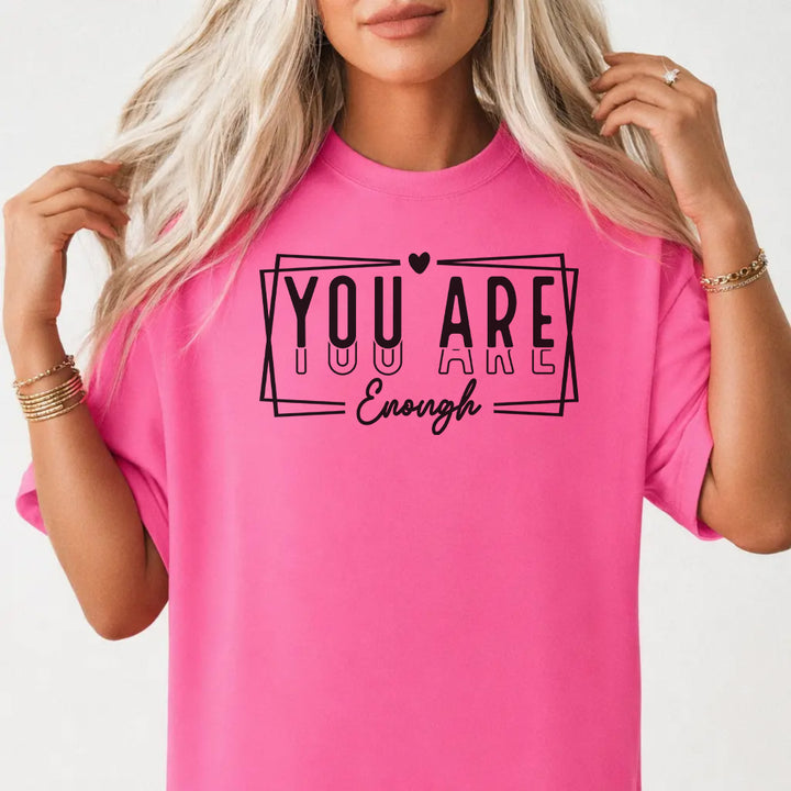 Woman wearing a neon pink graphic tee with bold stacked lettering and a heart accent that reads You Are Enough on the front