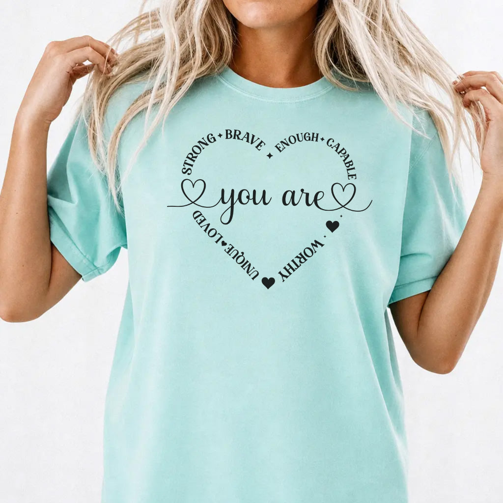 Woman wearing a chalky mint graphic tee with a heart made of affirmation words including strong brave worthy and loved around the phrase You Are on the front