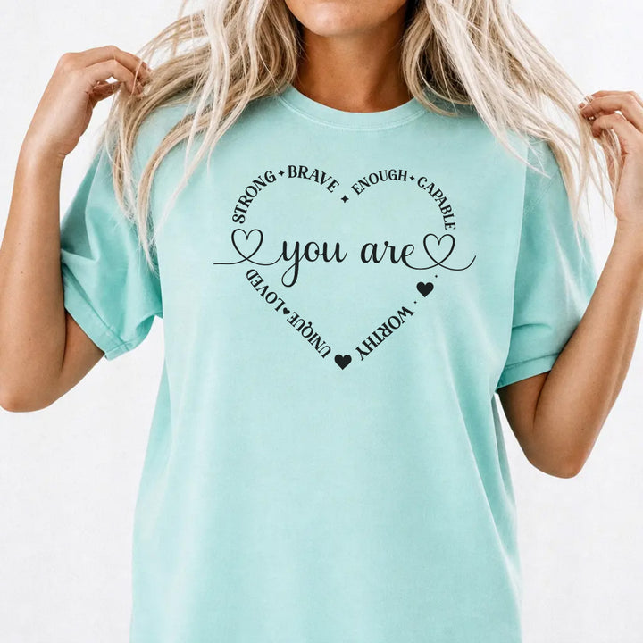 Woman wearing a chalky mint graphic tee with a heart made of affirmation words including strong brave worthy and loved around the phrase You Are on the front