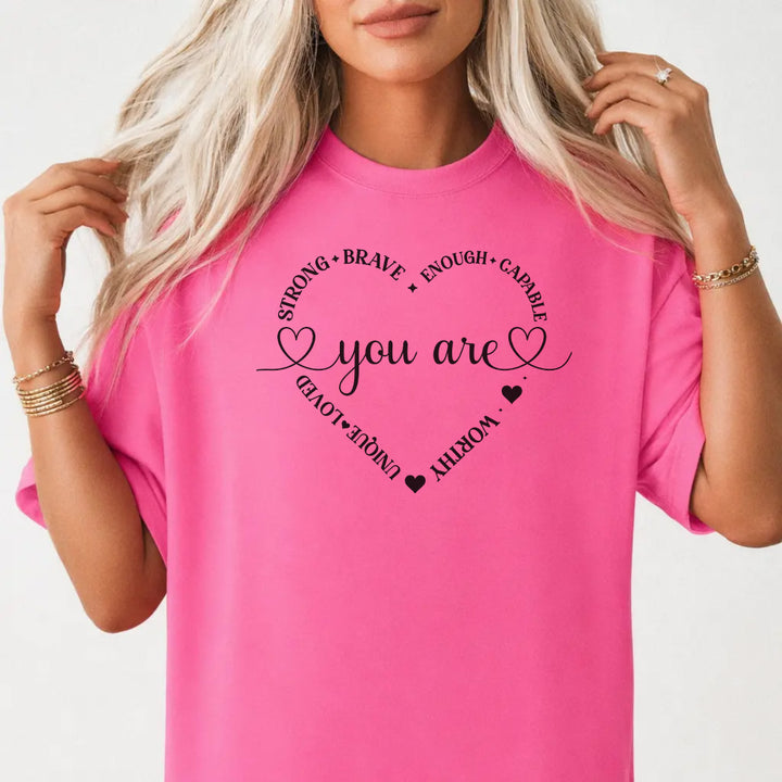 Woman wearing a neon pink graphic tee with a heart made of affirmation words including strong brave worthy and loved around the phrase You Are on the front