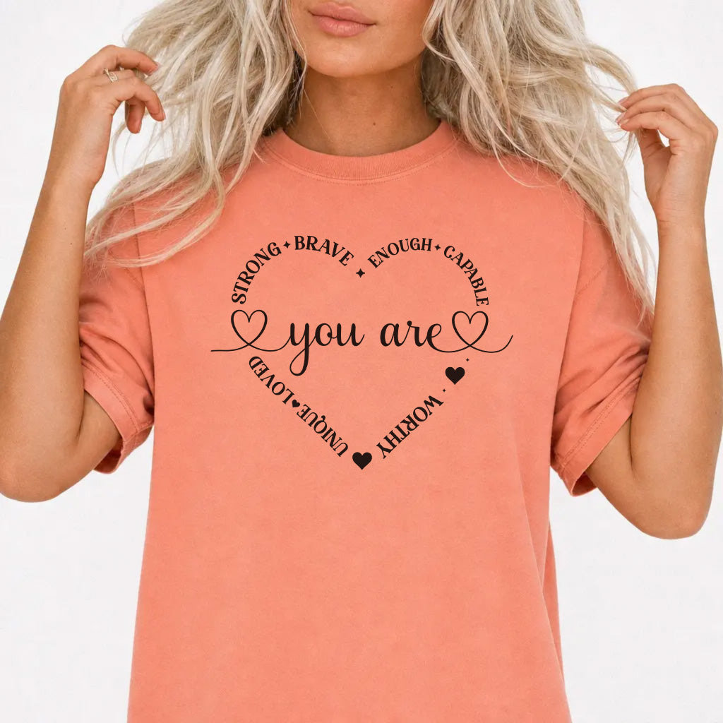 Woman wearing a terracotta graphic tee with a heart made of affirmation words including strong brave worthy and loved around the phrase You Are on the front