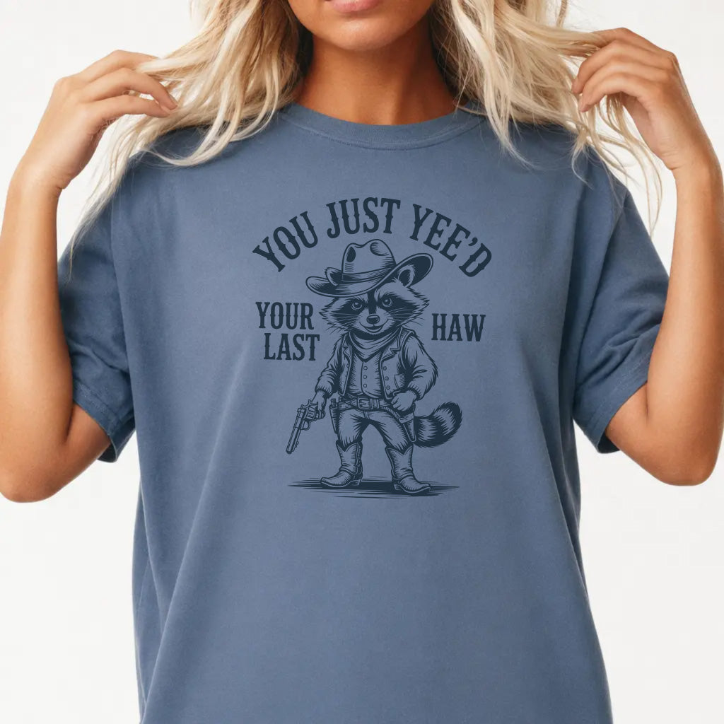 Woman wearing a blue jean graphic tee with a raccoon cowboy illustration and the phrase You Just Yeed Your Last Haw on the front