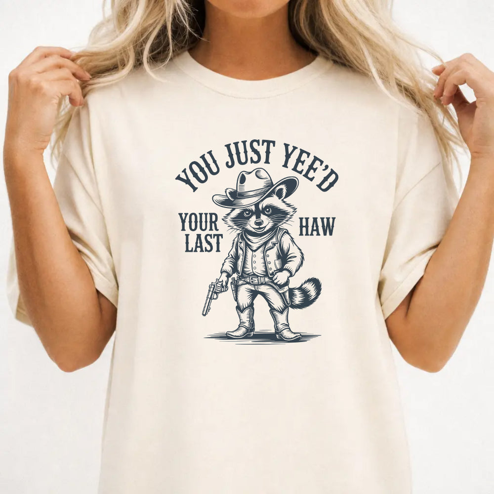 Woman wearing an ivory graphic tee with a raccoon cowboy illustration and the phrase You Just Yeed Your Last Haw on the front