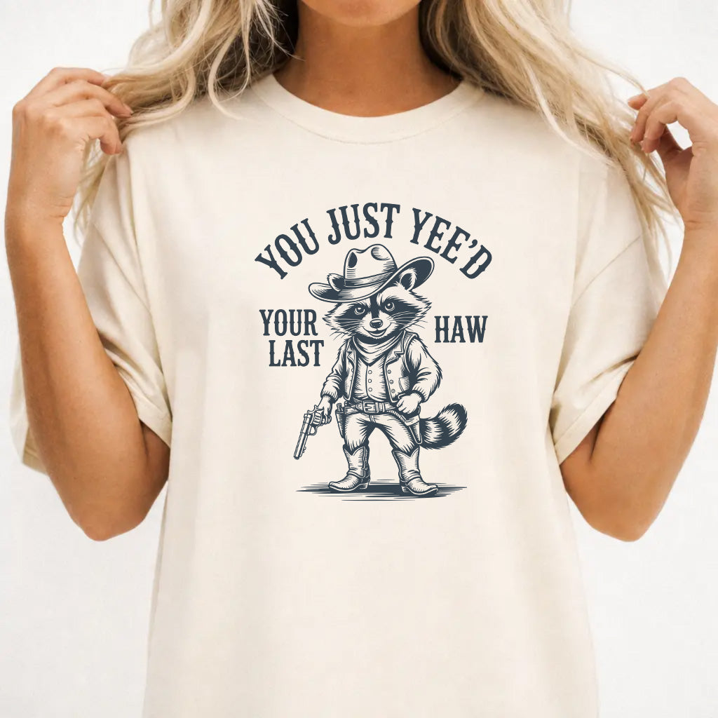 Woman wearing an ivory graphic tee with a raccoon cowboy illustration and the phrase You Just Yeed Your Last Haw on the front