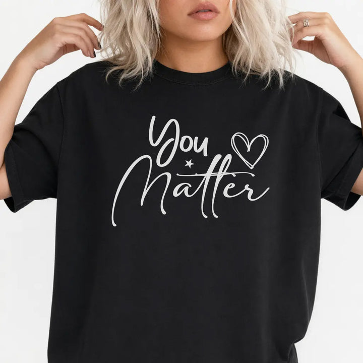 Woman wearing a black graphic tee with white script lettering and a hand-drawn heart that reads You Matter on the front