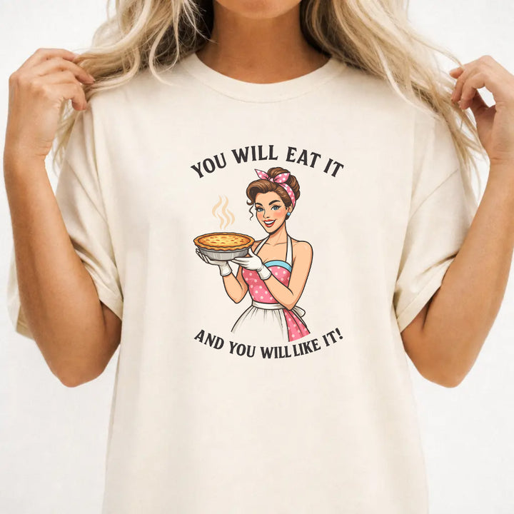 Woman wearing an ivory graphic tee with a retro pin-up woman holding a pie and the phrase You Will Eat It and You Will Like It on the front