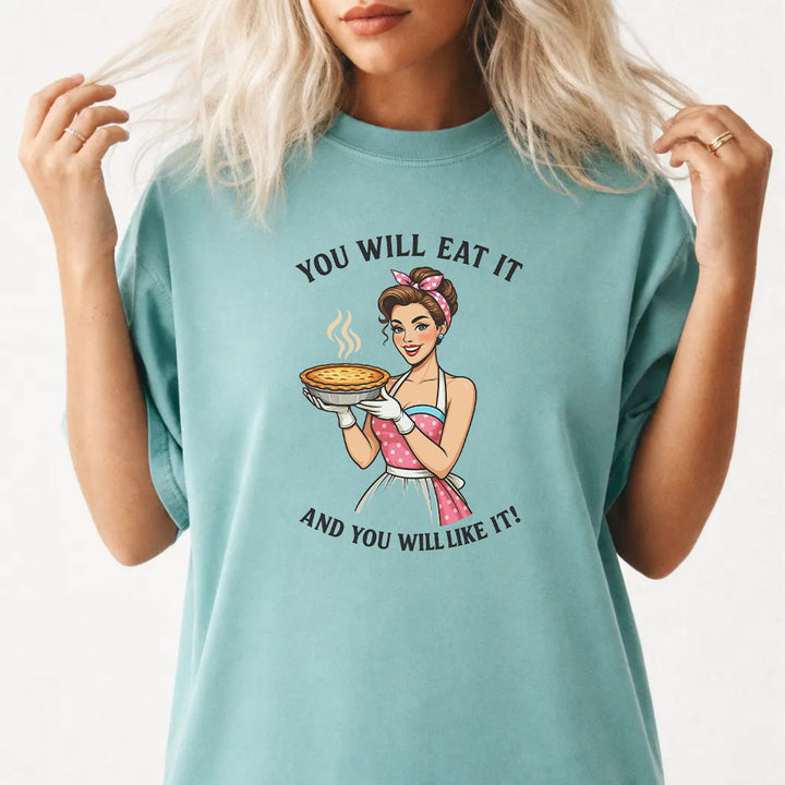 Woman wearing a seafoam graphic tee with a retro pin-up woman holding a pie and the phrase You Will Eat It and You Will Like It on the front